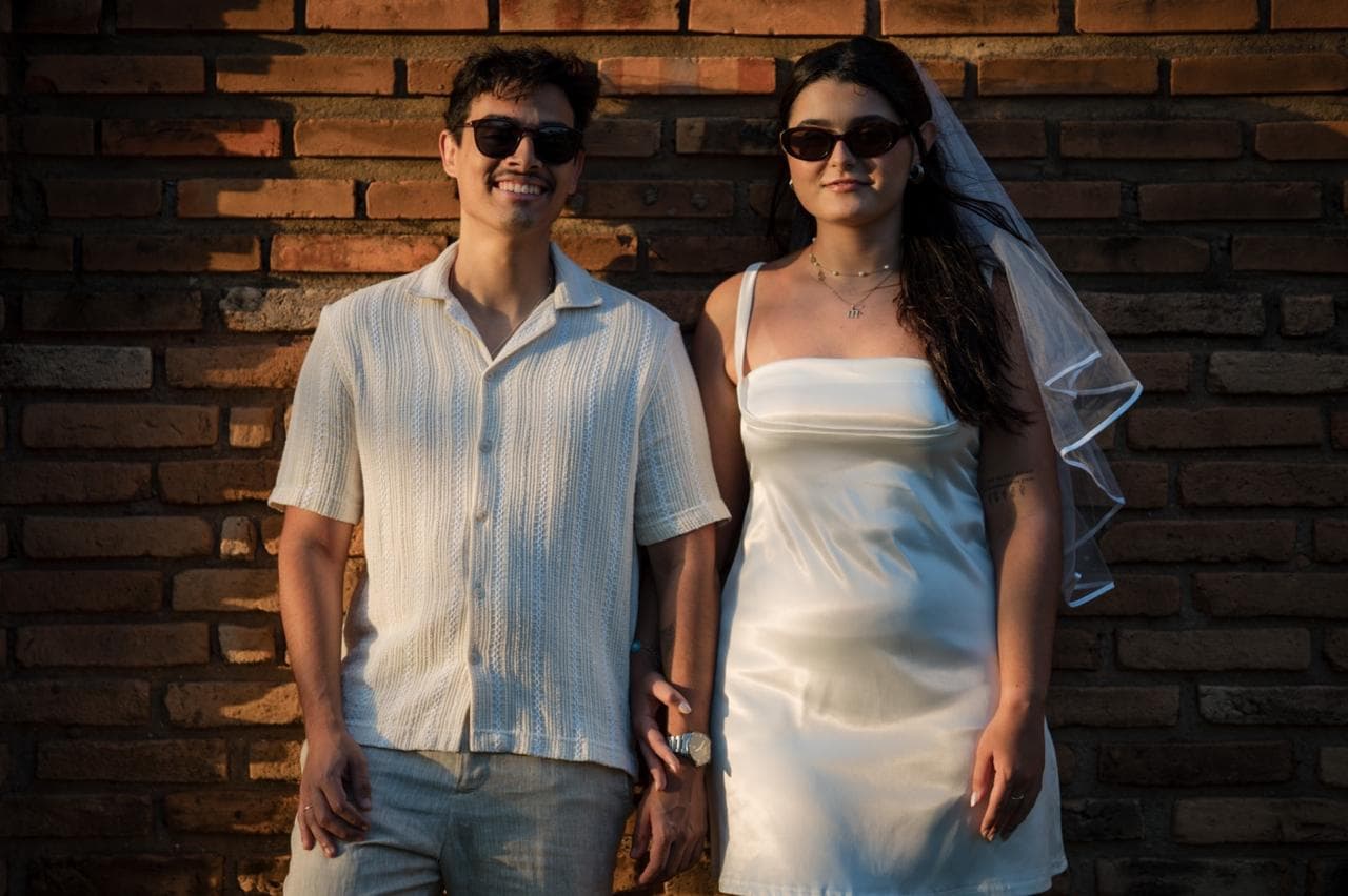 Lucas e Marina vestidos de noivos em frente a um muro de tijolos
