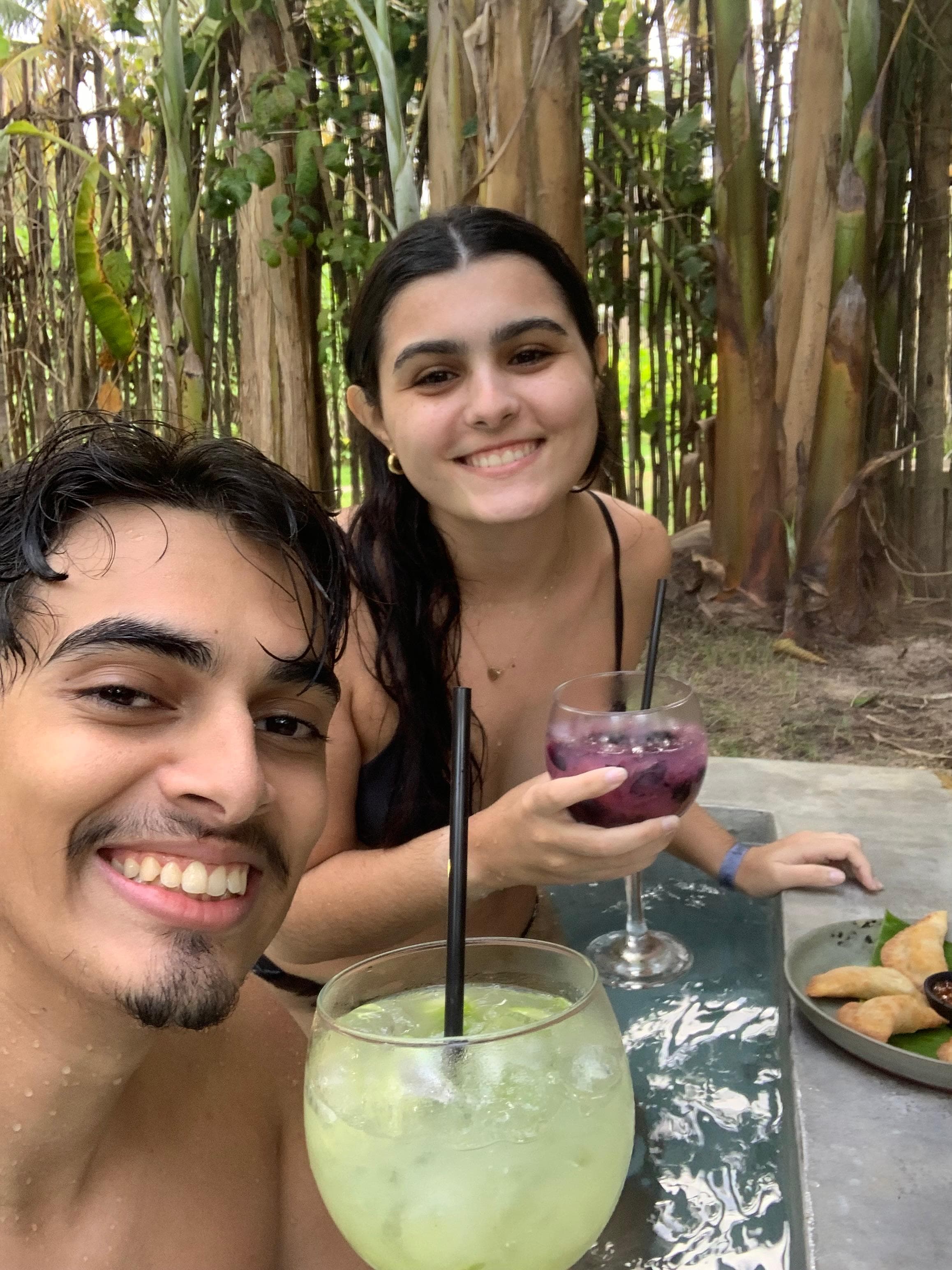 Lucas e Marina com drinks em um bar à beira-mar entre coqueiros